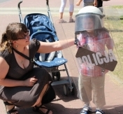 Na zdjęciu dzieco w kasku i z tarczą policyjną. Obok kuca mama. W tle wózek dziecięcy. Na zdjęciu dzieco w kasku i z tarczą policyjną. Obok kuca mama. W tle wózek dziecięcy.