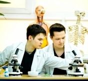 Dwóch uczniów w kitlach siedzi w pracowni biologicznej sprawdzją mikroskop.