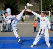 Dwóch zawodników przeprowadza pokaz sportowy. Jeden z nich trzyma deski na wysokości głowy. Drugi uderza z całej siły w nią stopą.zdj. Krzysztof Dobrogowski (http://csp.edu.pl Centrum Szkolenia Policji)