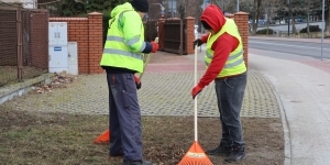 dwaj mężczyźni w odblaskowych kamizelkach grabią liście