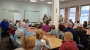 Jedna osoba stoi, kilkanaście osób siedzi