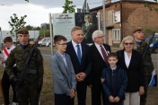 Kilka osób przed tablicą pamiątkową, w tym dwóch mężczyzn wo wojskowych mundurach i bronią.