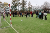 Grupa osób na boisku, chłopiec kopie piłkę do bramki