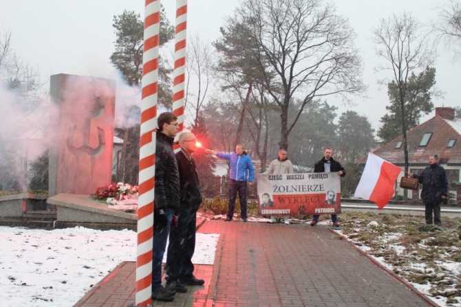 Grupa młodzieży pod pomnikiem. Dwie osoby trzymają rozłożoną flagę Polski. Z boku chłopak trzyma w ręku rozpaloną racę.