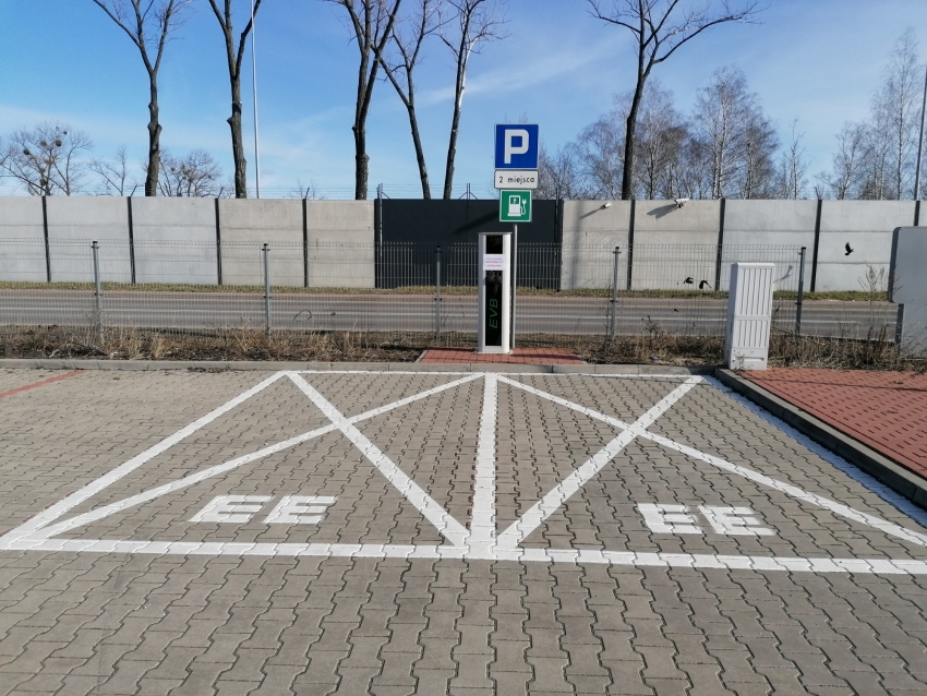 Na pierwszym planie znak - Parkuj i Jedź, w tle stacja do ładowania samochodów elektrycznych.