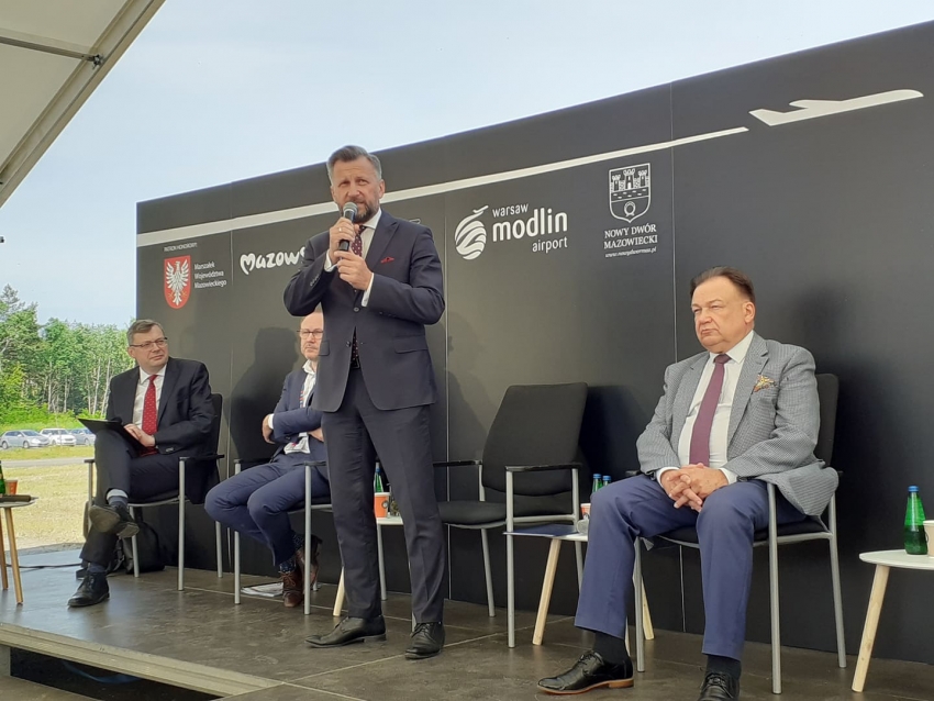Cztery osoby na tle ścianki, jedne z nich stoi i mówi przez mikrofon, trzech siedzi.