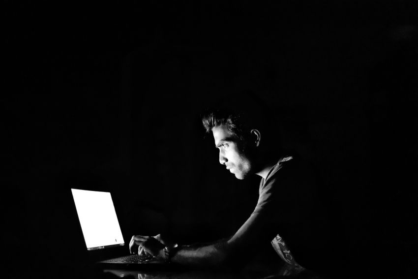 Na zdjęciu mężczyzna w ciemnościach piszący na laptopie