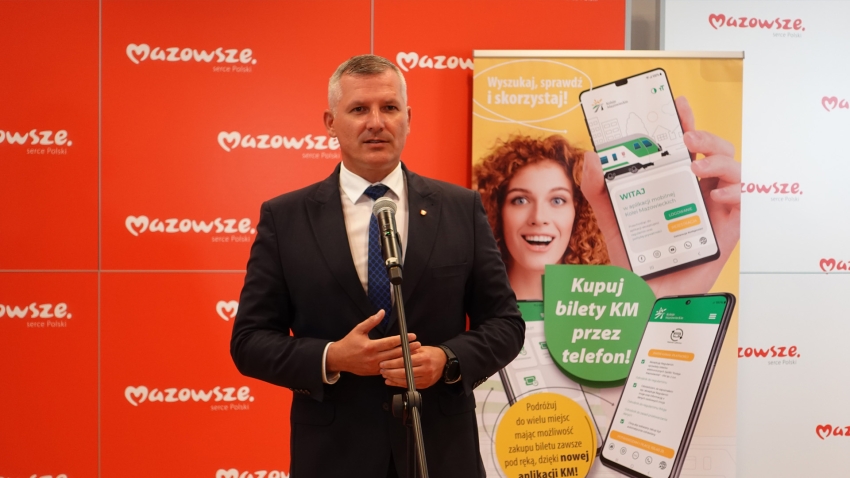 Mężczyzna przemawiający przez mikrofon, w tle reklama aplikacji do kupowania biletów Kolei Mazowieckich
