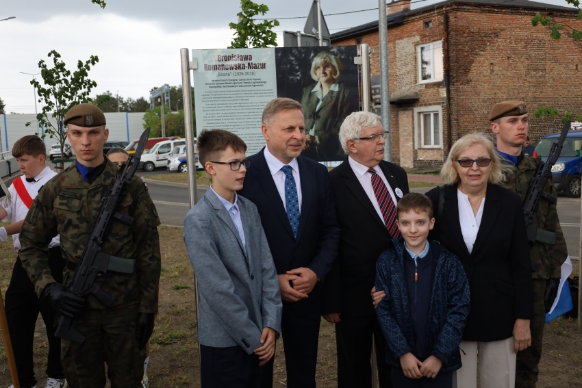 Kilka osób przed tablicą pamiątkową, w tym dwóch mężczyzn wo wojskowych mundurach i bronią.