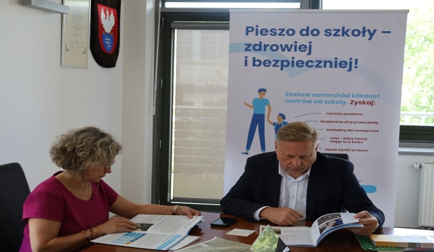 dwie osoby siedzą przy biurku i czytają kalatogi, z tyłu stoi plakat z napisem pieszo do szkoły - zdrowiej i bezpieczniej