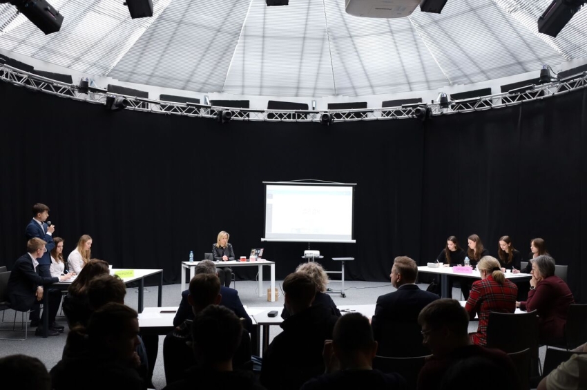 młodzież zebrana w sali w czasie debaty. dwie drużyny siedzą przy dwóch stolikach, na przeciwko nich siedzi jury a za nimi widownia