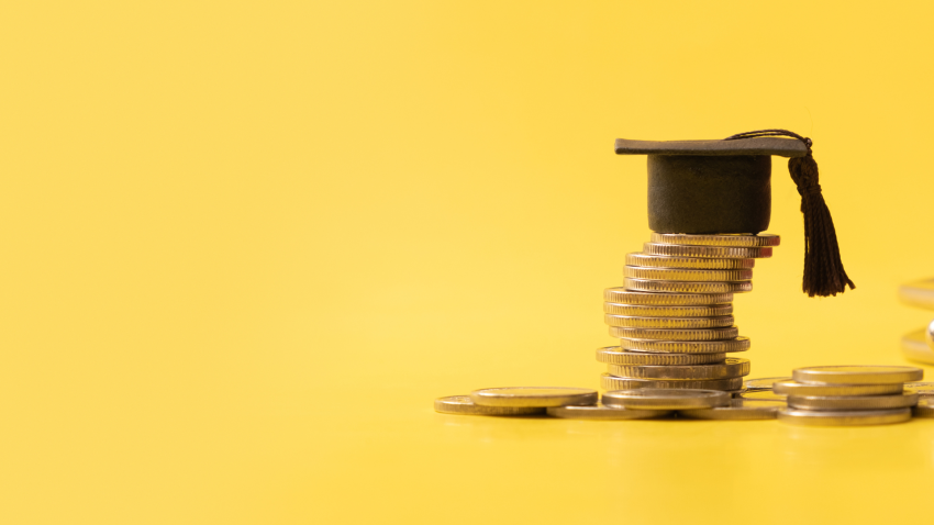 żółte tło, monety ułożone w stos, na wierzchu studencka czapka